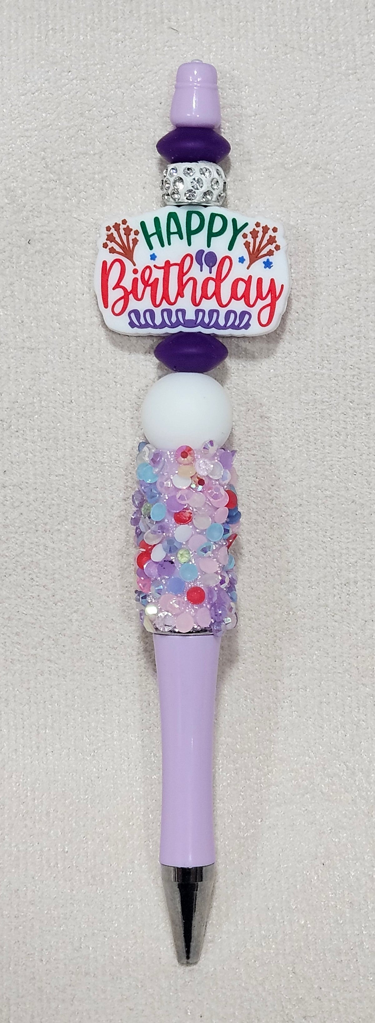 A custom purple and white pen decorated with a 'Happy Birthday' sign, a purple cap, a white bead, a glittery rhinestone grip, and a silver metal tip.